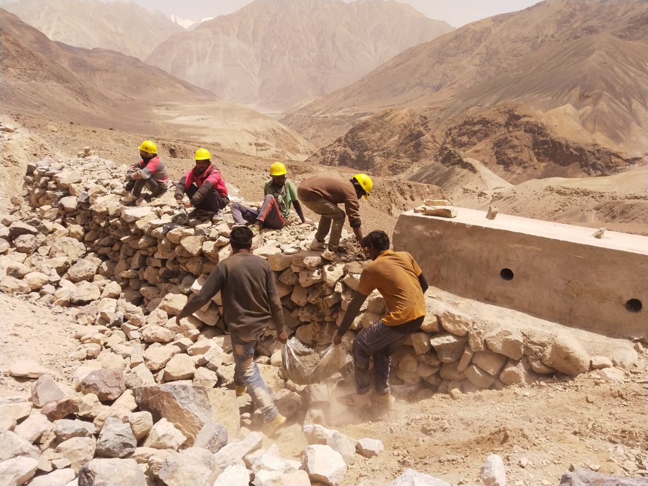 Road labourers building a stone retaining wall in a Ladakh valley, dust and rubble around them.