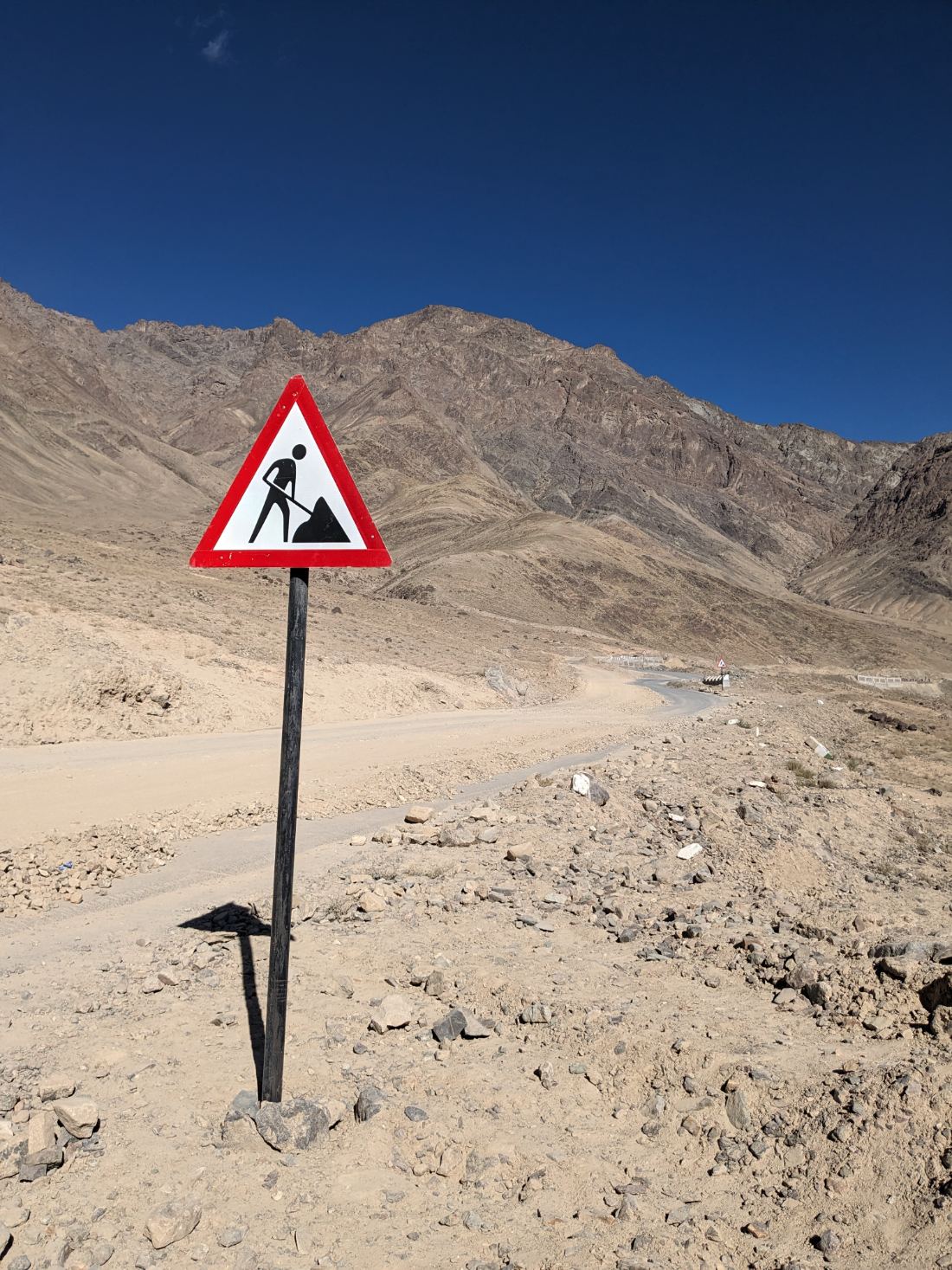 A red triangular men-at-work sign on a wooden post against a blue Ladakh sky and rocky mountain backdrop.