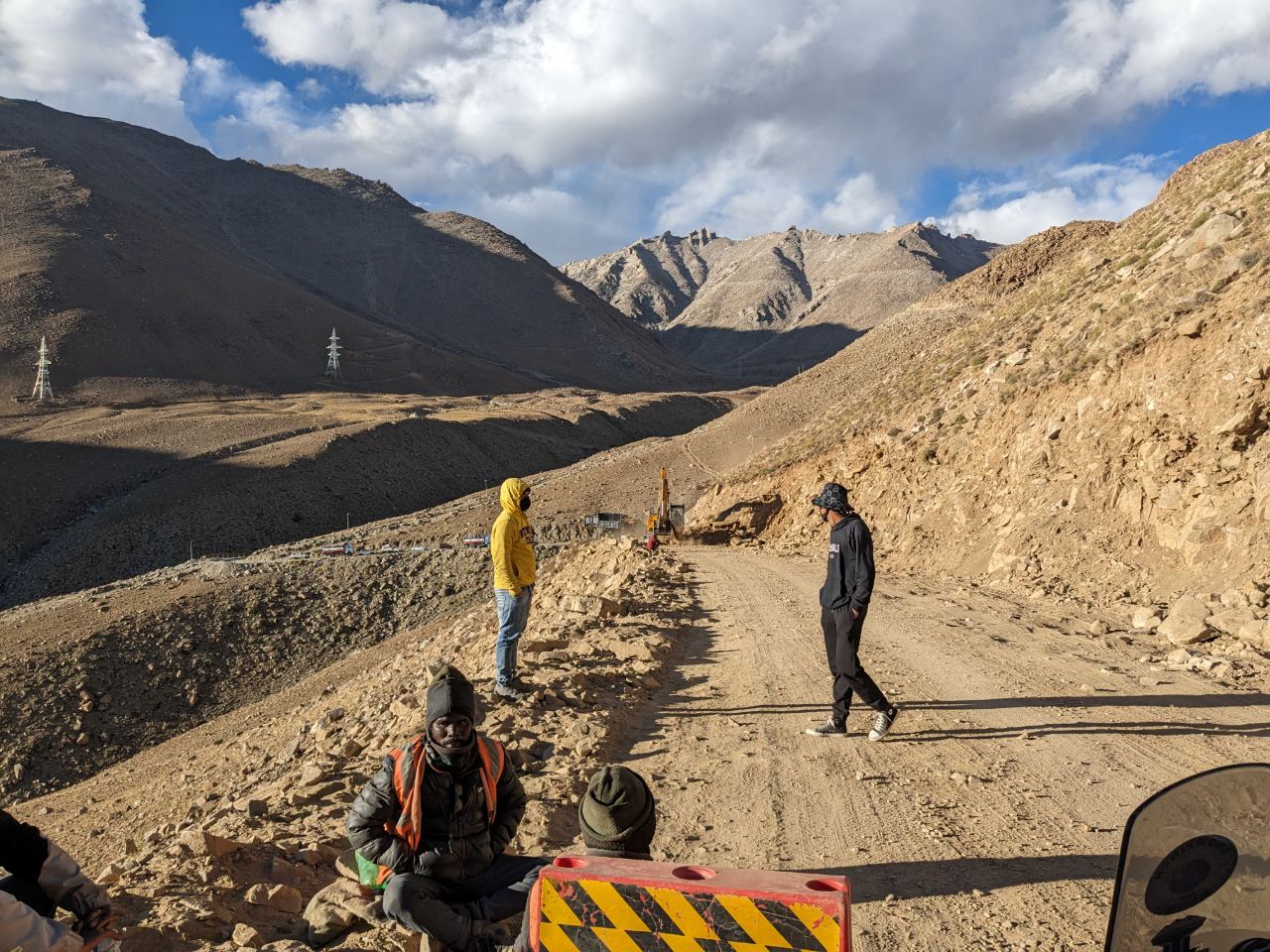 Road construction crew at altitude along a Ladakh valley — workers resting beside a striped barrier.